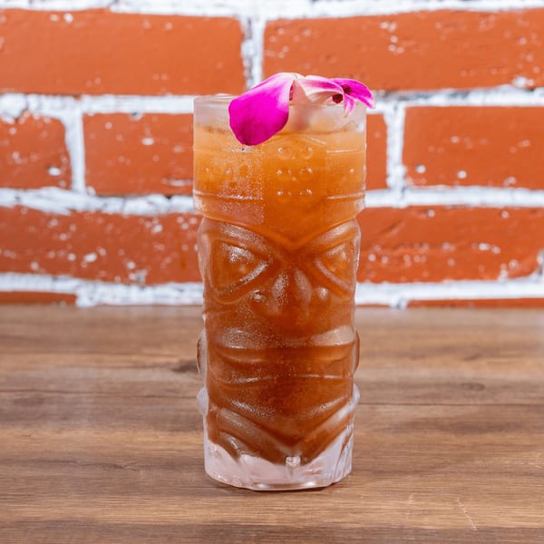 A tiki-style glass filled with a brown beverage and garnished with a pink orchid.