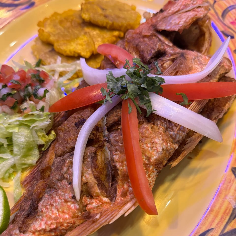 Whole fried fish garnished with onions, tomatoes, and cilantro, served with plantains and salad.