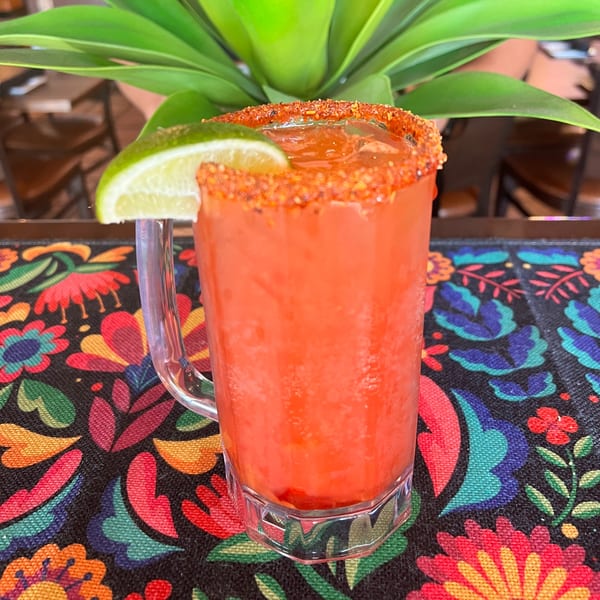 A red Mexican drink with a chili-lime rim and a lime wedge, on a colorful floral placemat.
