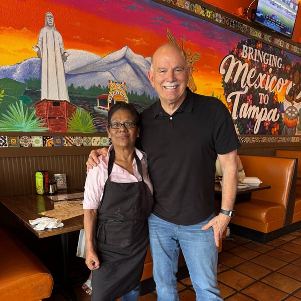 Two people, a woman in an apron and a man, smiling in front of a vibrant Mexican mural.
